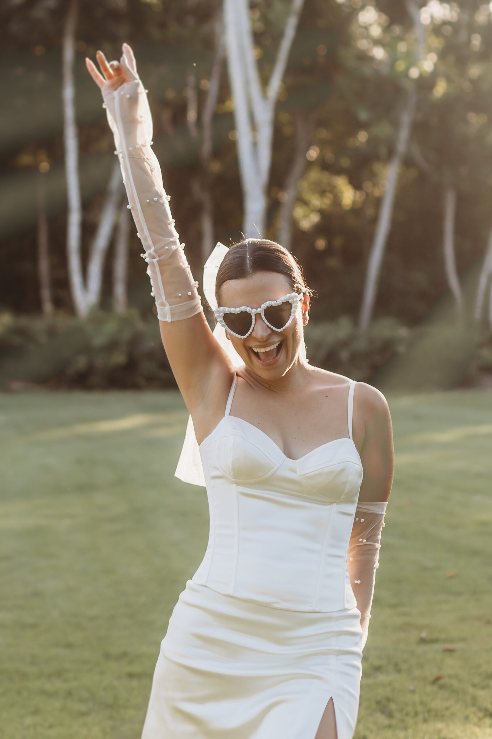 A fun and stylish bride with heart-shaped pearl sunglasses and mesh pearl gloves raises her hand in celebration in a lush La Romana garden, perfect for a destination wedding in the Dominican Republic. Photographed by a La Romana Photographer.