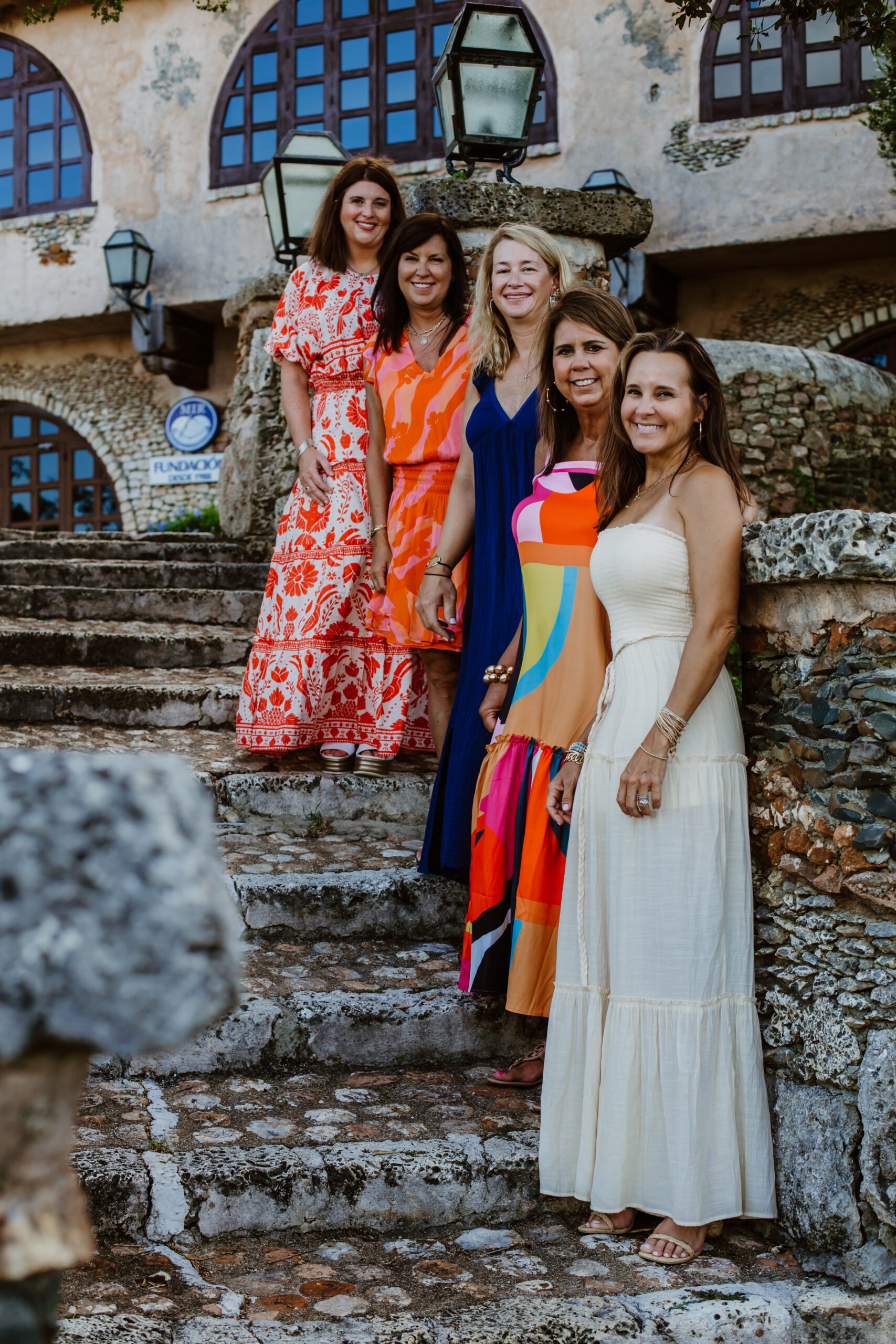 Five happy women in colorful dresses, including a cream-white maxi, a patterned red-and-white, and an orange-and-white print, pose on historic stone steps during a group photoshoot with a La Romana photographer in the Dominican Republic. The background features ancient stone architecture and arched windows.