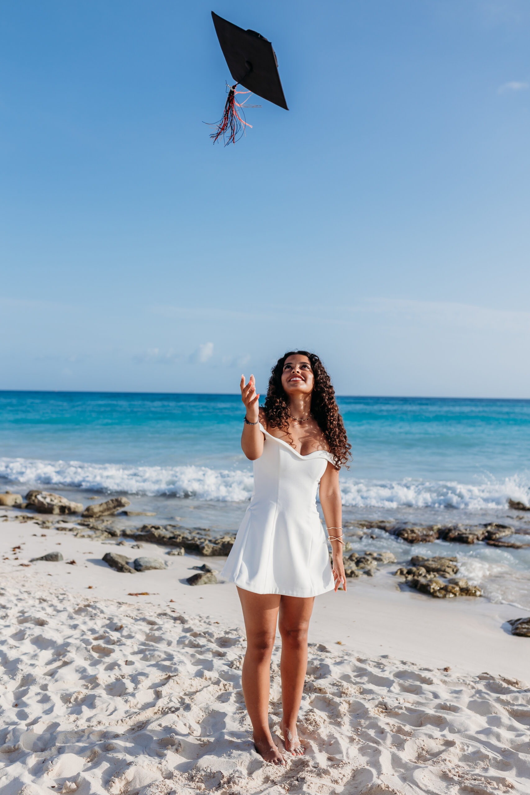 A joyful young female graduate with long curly hair, wearing a white off-the-shoulder dress, throws her black graduation cap into the clear blue sky with excitement on a tropical beach in La Romana, Dominican Republic, captured by a professional La Romana photographer.