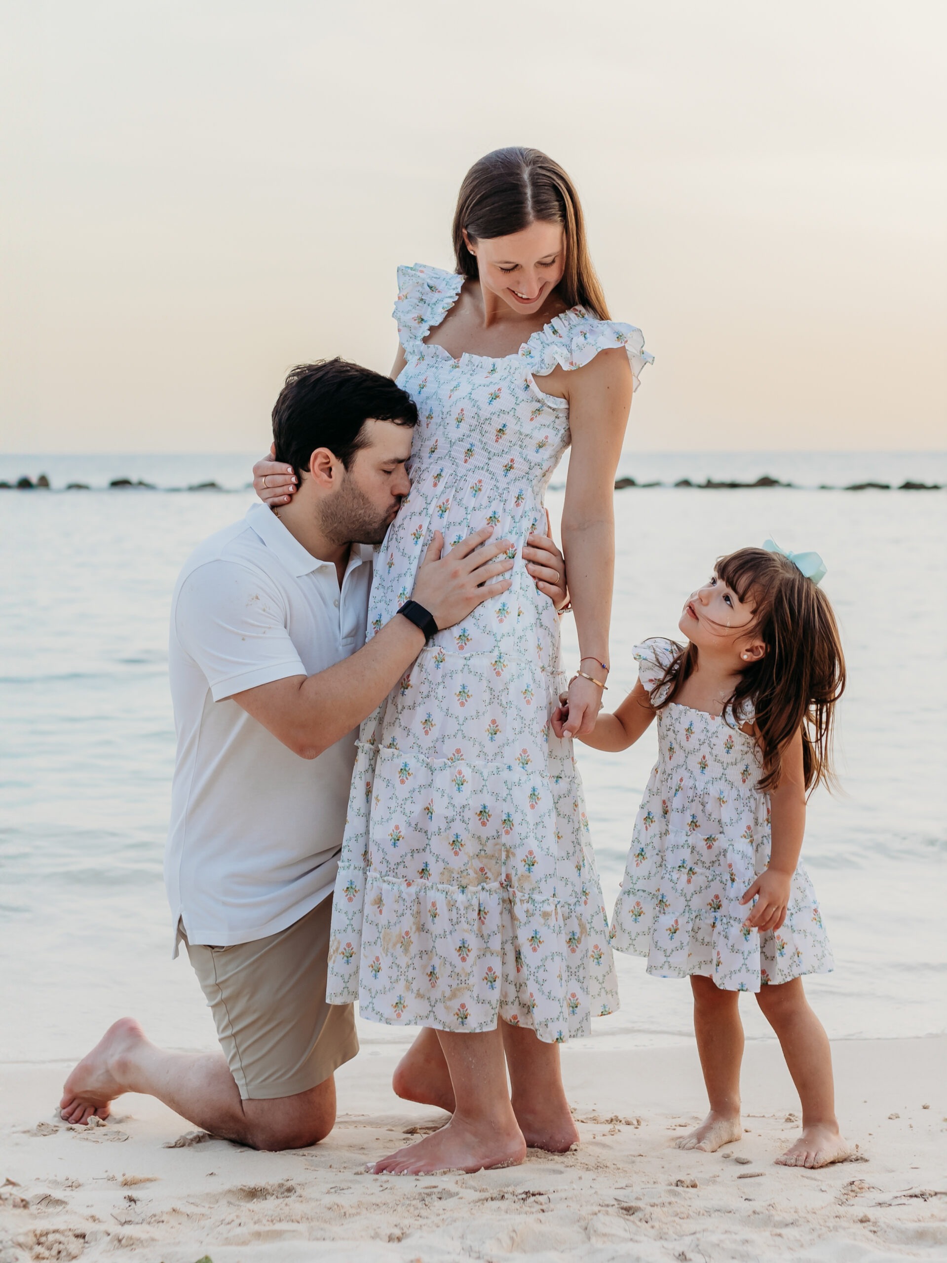 Family maternity photoshoot in La Romana by photographer at Casa de Campo, showing a husband kissing his pregnant wife's bump while their young daughter stands beside them on Minitas Beach