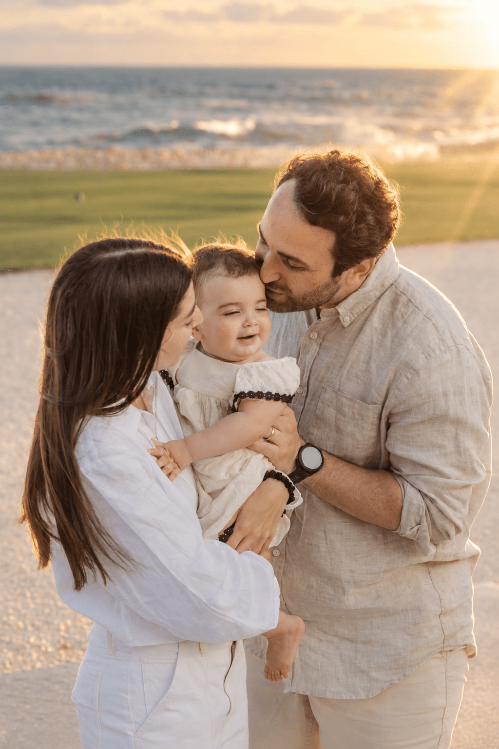 Loving parents holding and kissing their baby daughter during a golden hour sunset family photoshoot at Punta Espada Golf Club in Cap Cana, captured by a La Romana photographer.
