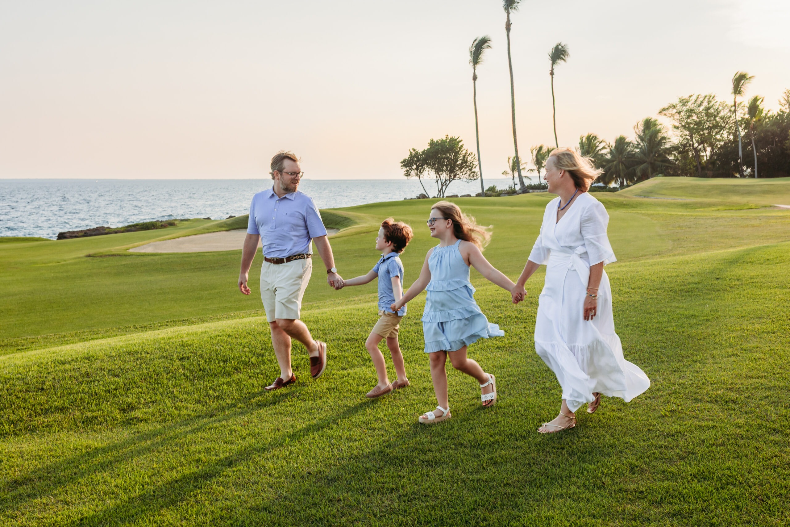 Family of four holding hands and walking across a lush green oceanfront golf course at golden hour in Cap Cana, captured during a resort family portrait session by a La Romana photographer.