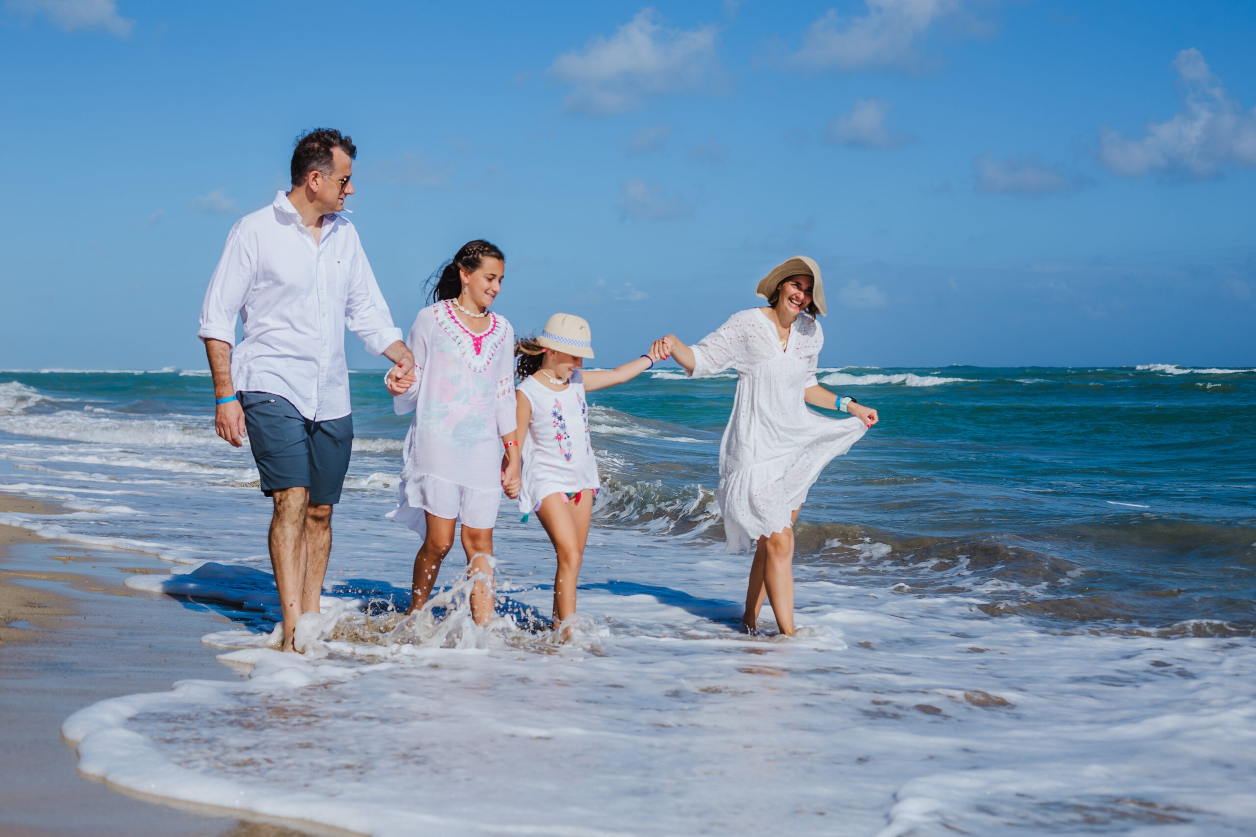 Happy family of four walking hand in hand through shallow ocean waves on the beach in Miches during a tropical vacation photoshoot, captured by a La Romana photographer.