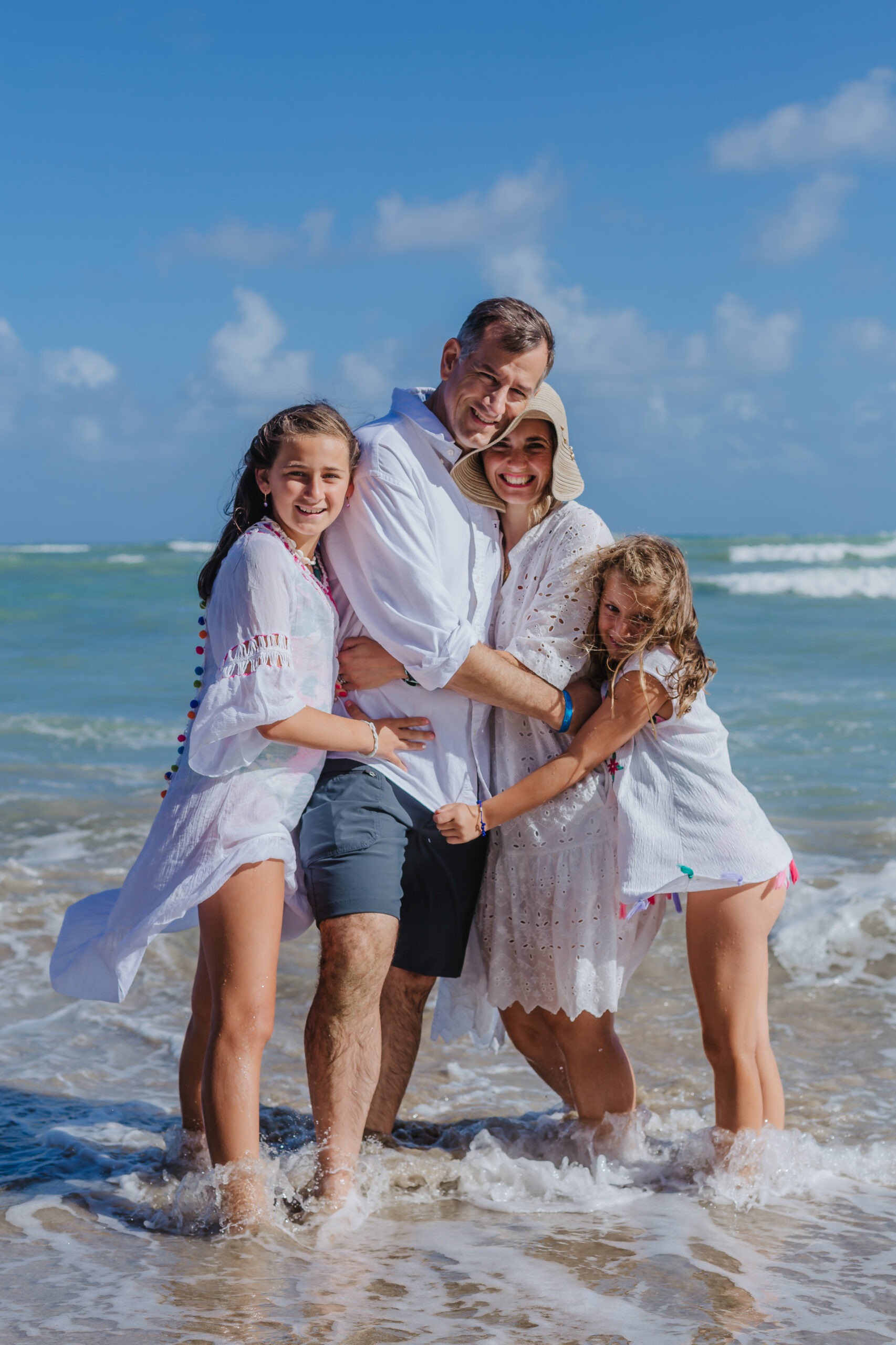 Happy family of four hugging and smiling while standing in shallow ocean waves during a beach vacation photoshoot, captured by a La Romana photographer in the Dominican Republic.
