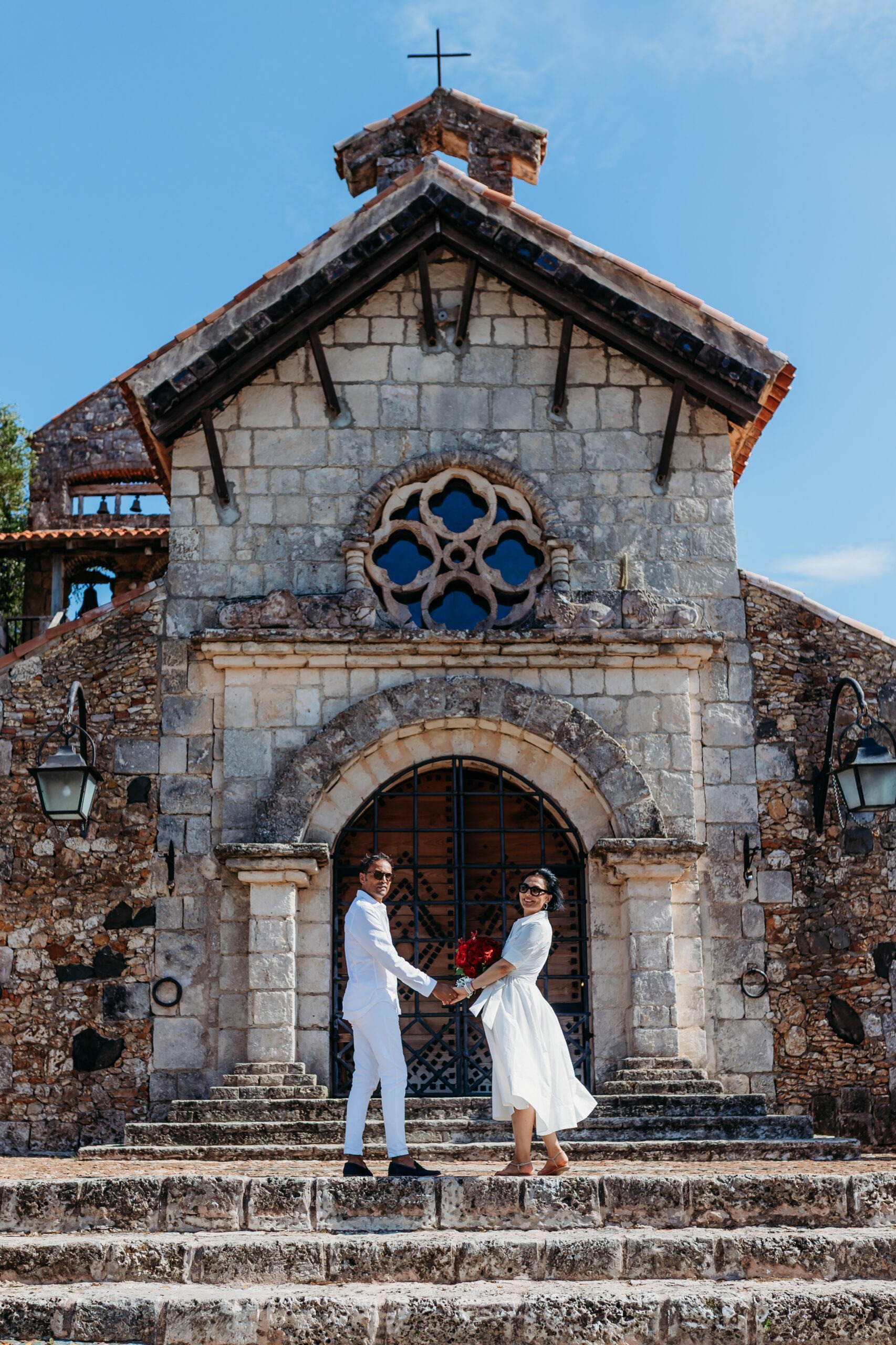 Couple dressed in white holding hands with a red rose bouquet outside the historic St. Stanislaus stone church in Altos de Chavón, Casa de Campo, during a vow renewal photoshoot with a La Romana photographer in the Dominican Republic.