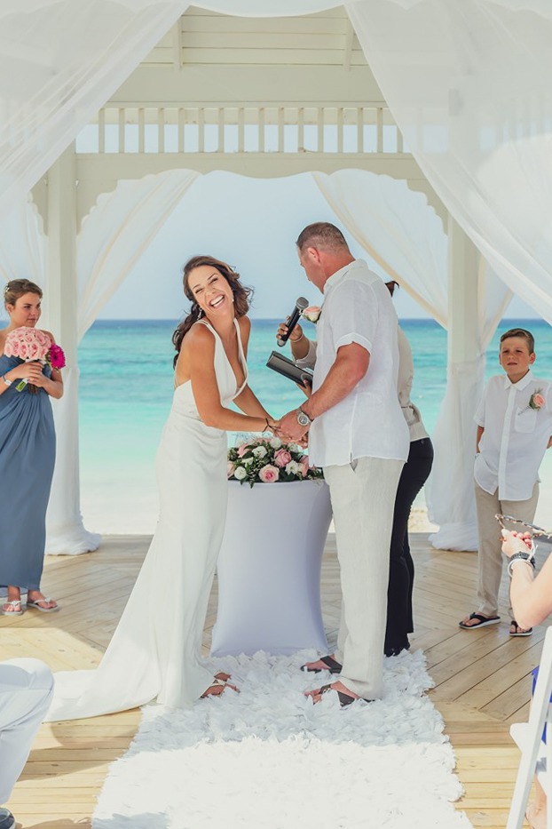 Joyful bride laughing during a beach vow renewal ceremony under a white draped gazebo overlooking turquoise ocean waters in Cap Cana, captured by a La Romana photographer in the Dominican Republic.